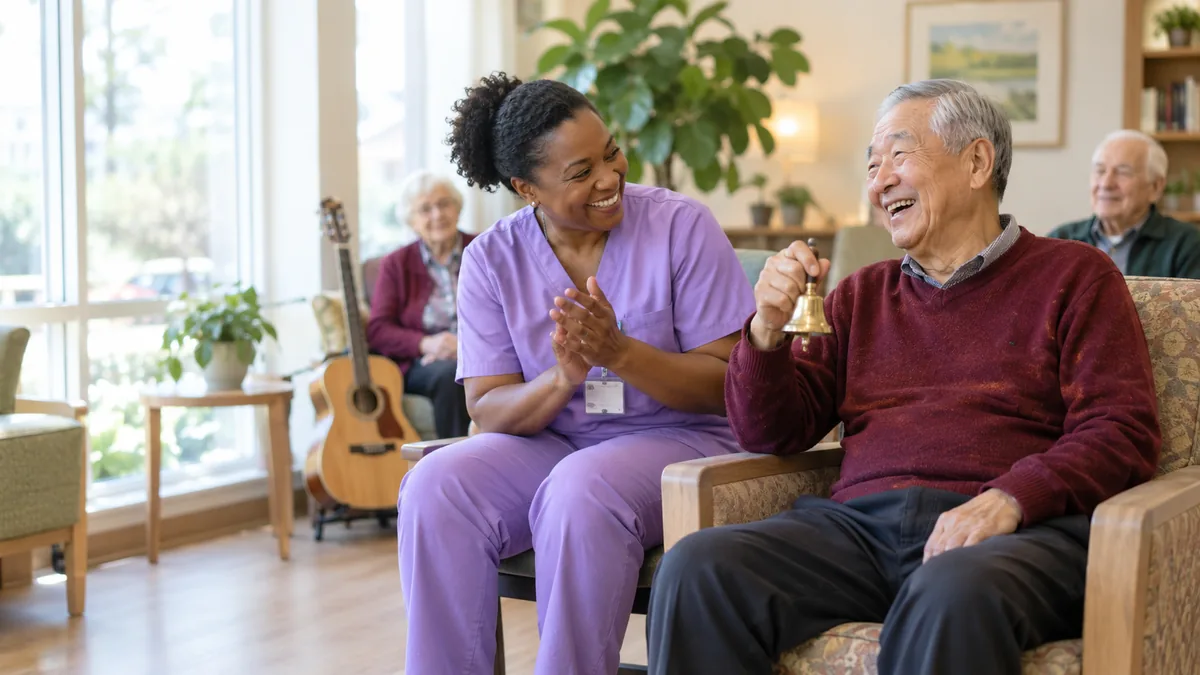 Older man shaking a hand bell and smiling with a caregiver during a music session in memory care